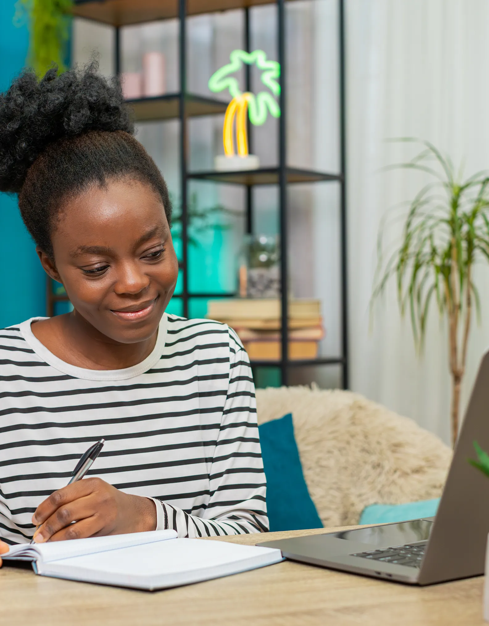 Woman in a striped shirt writes in a notebook at a desk with a laptop in a bright, modern workspace.