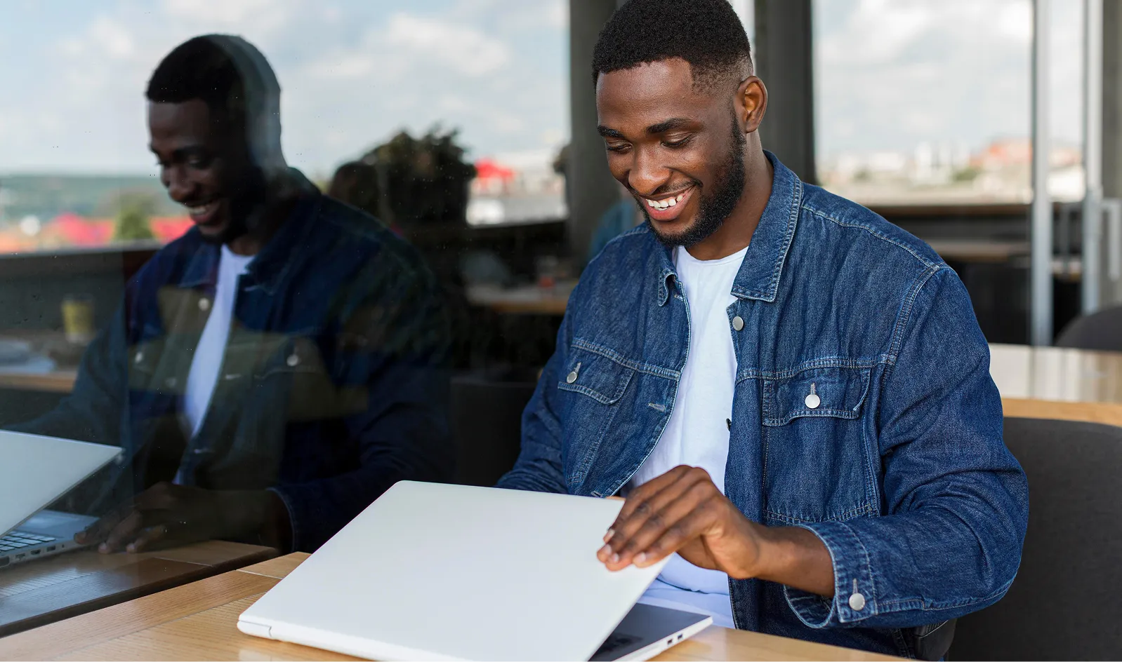 Young man in a denim jacket typing on a white laptop at a cafe, smiling while looking at the screen.