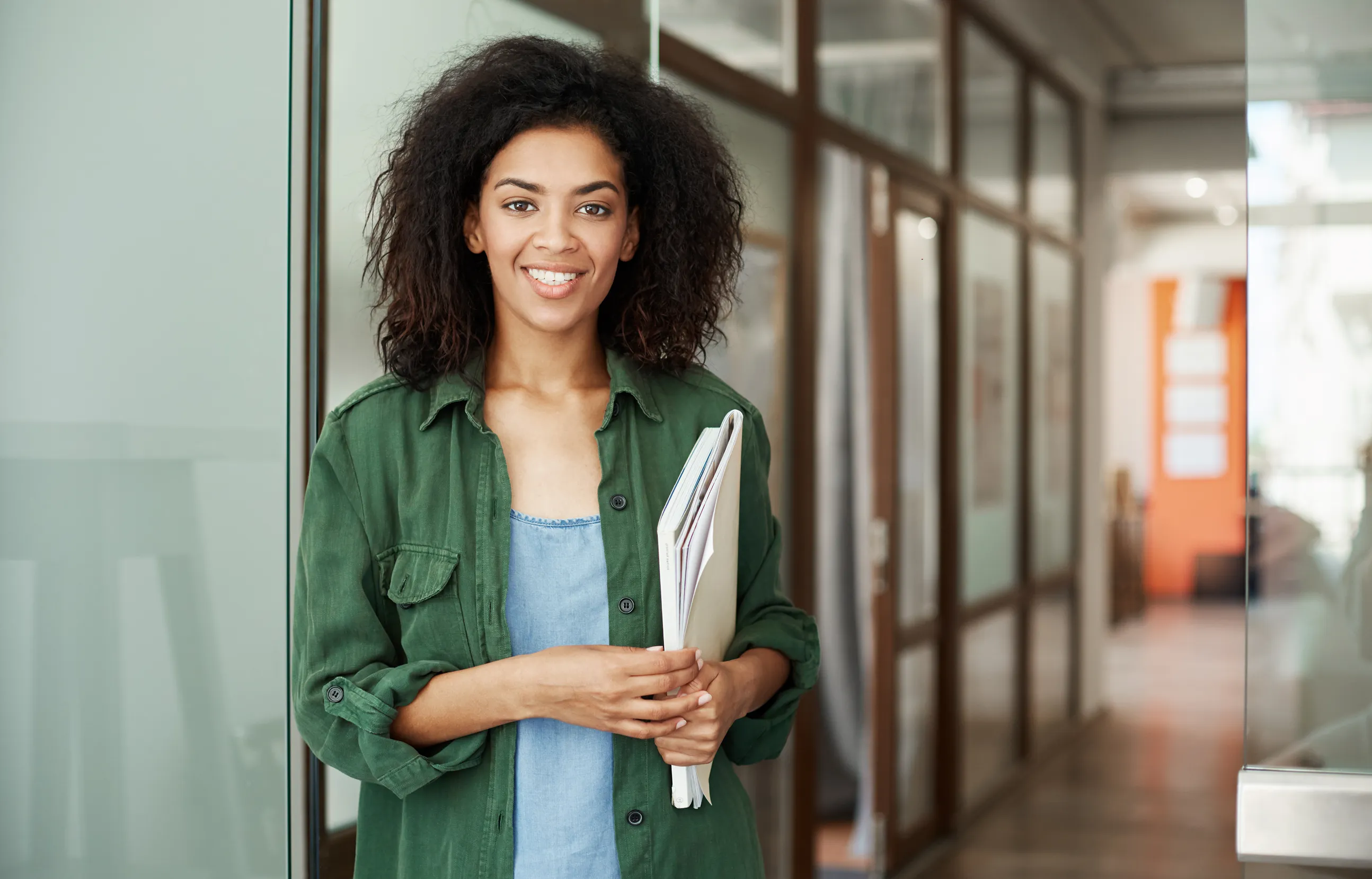Smiling woman with curly hair in a green shirt stands by a glass wall in a modern hallway, holding papers.