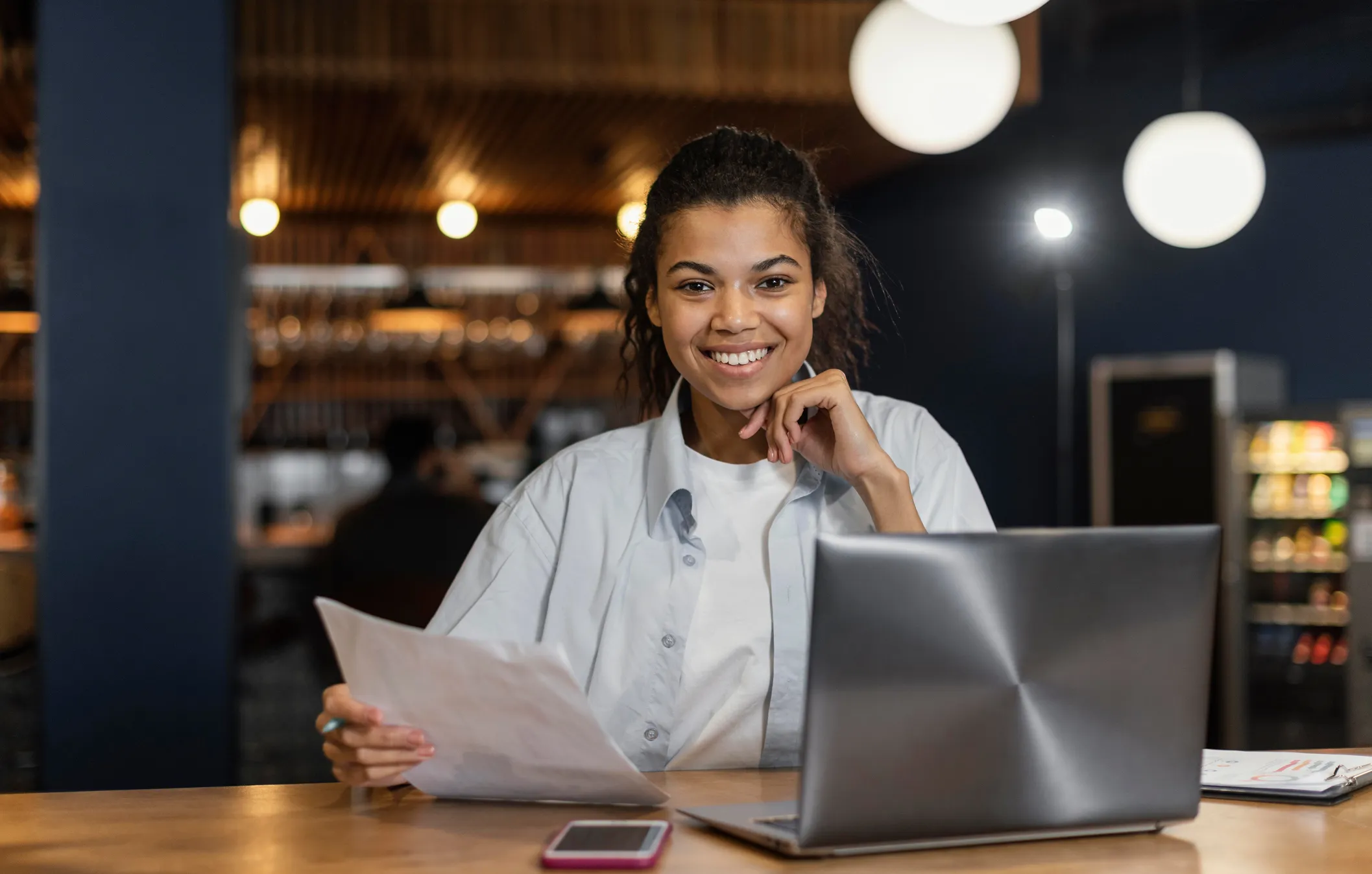 Smiling woman in a light shirt at a desk, reviewing a sheet of paper with a laptop and phone on the table.