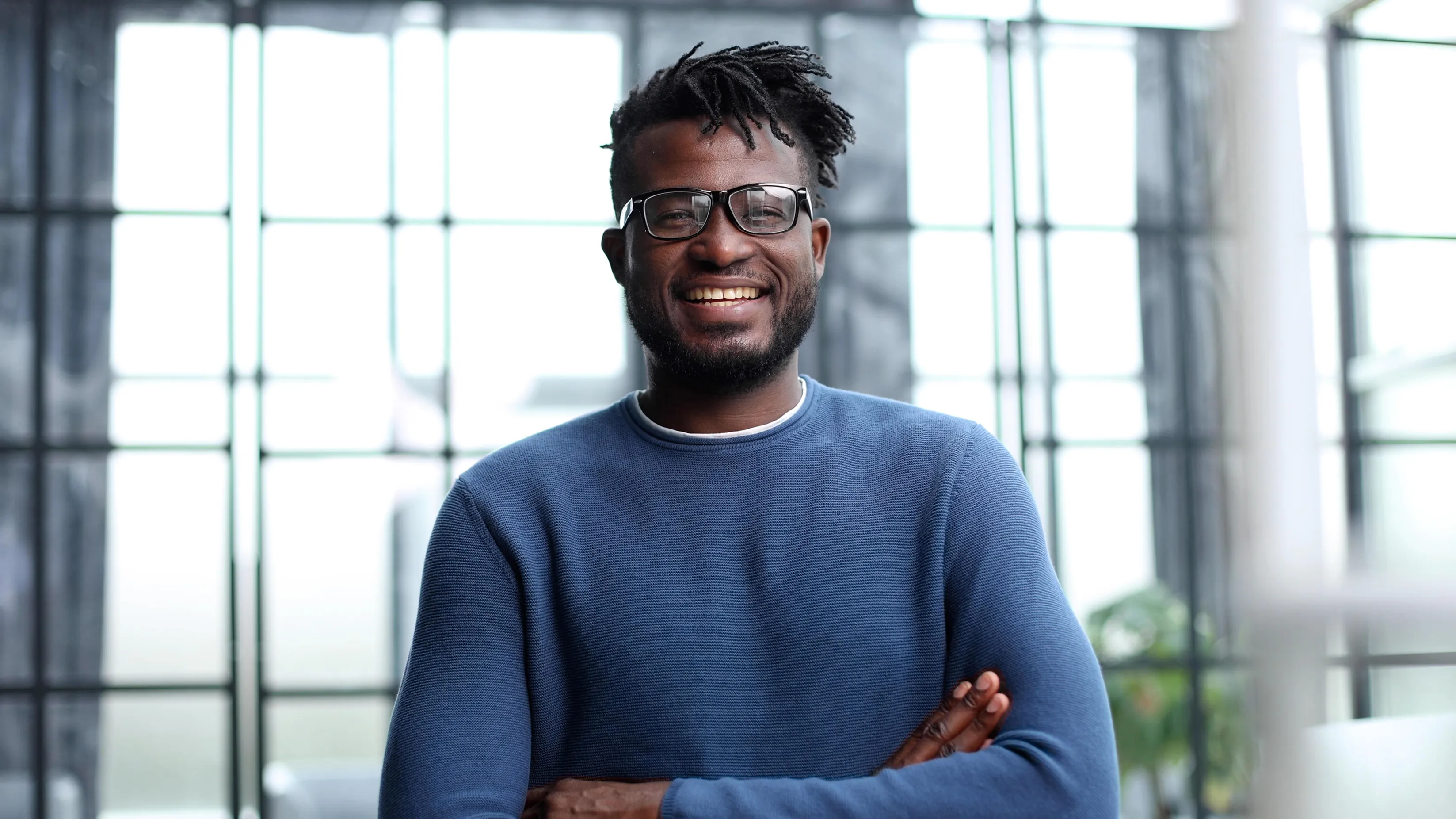Smiling man with glasses in a blue sweater, standing with arms crossed in a bright glass-walled space.