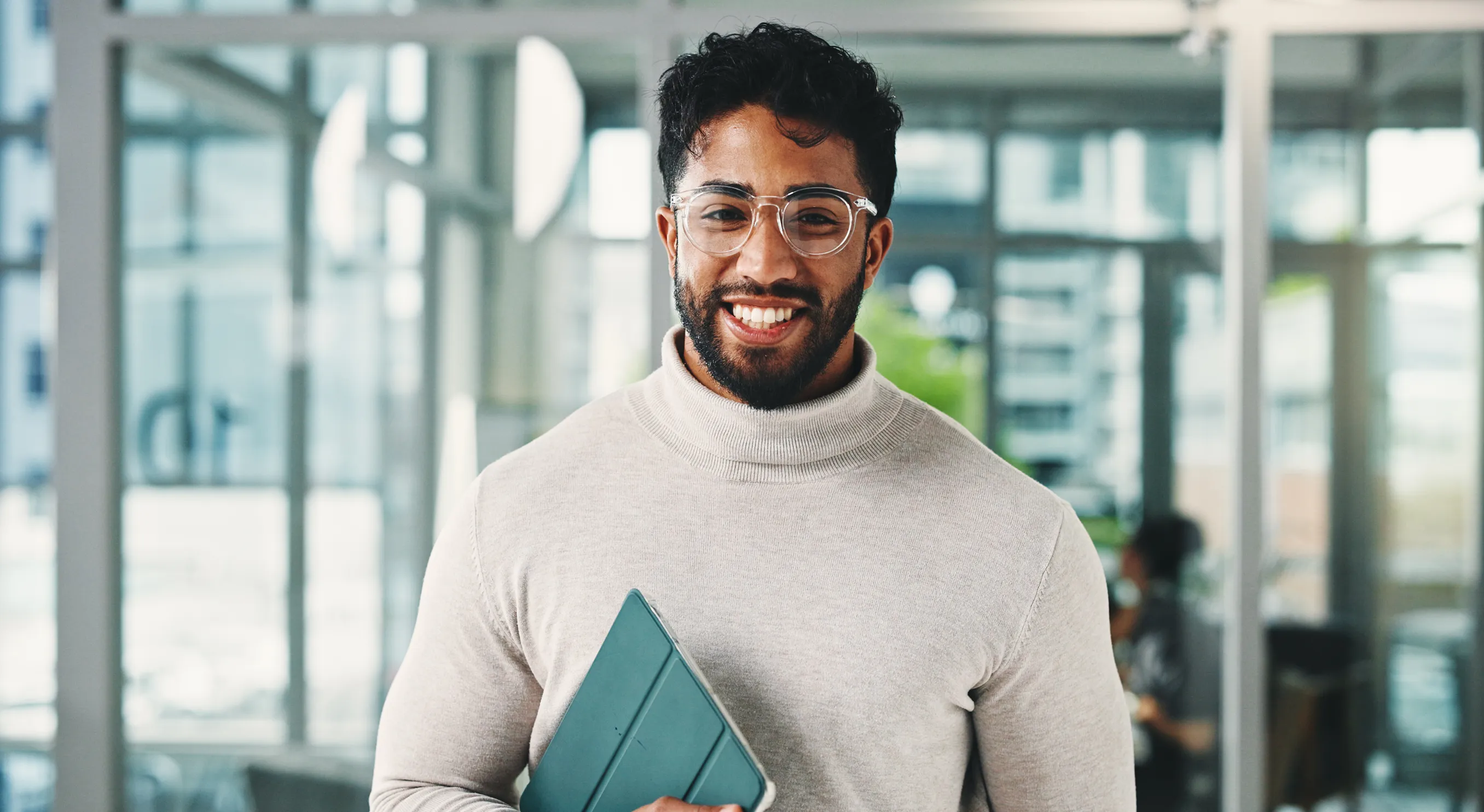 Smiling man with clear-frame glasses holding a teal binder in a bright office lobby