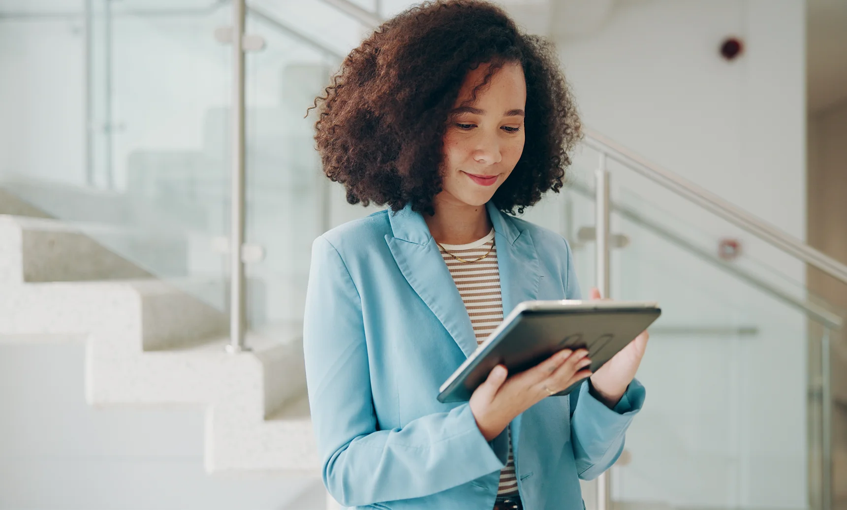 Professional woman in a light blue blazer reading a tablet in a bright, modern lobby.