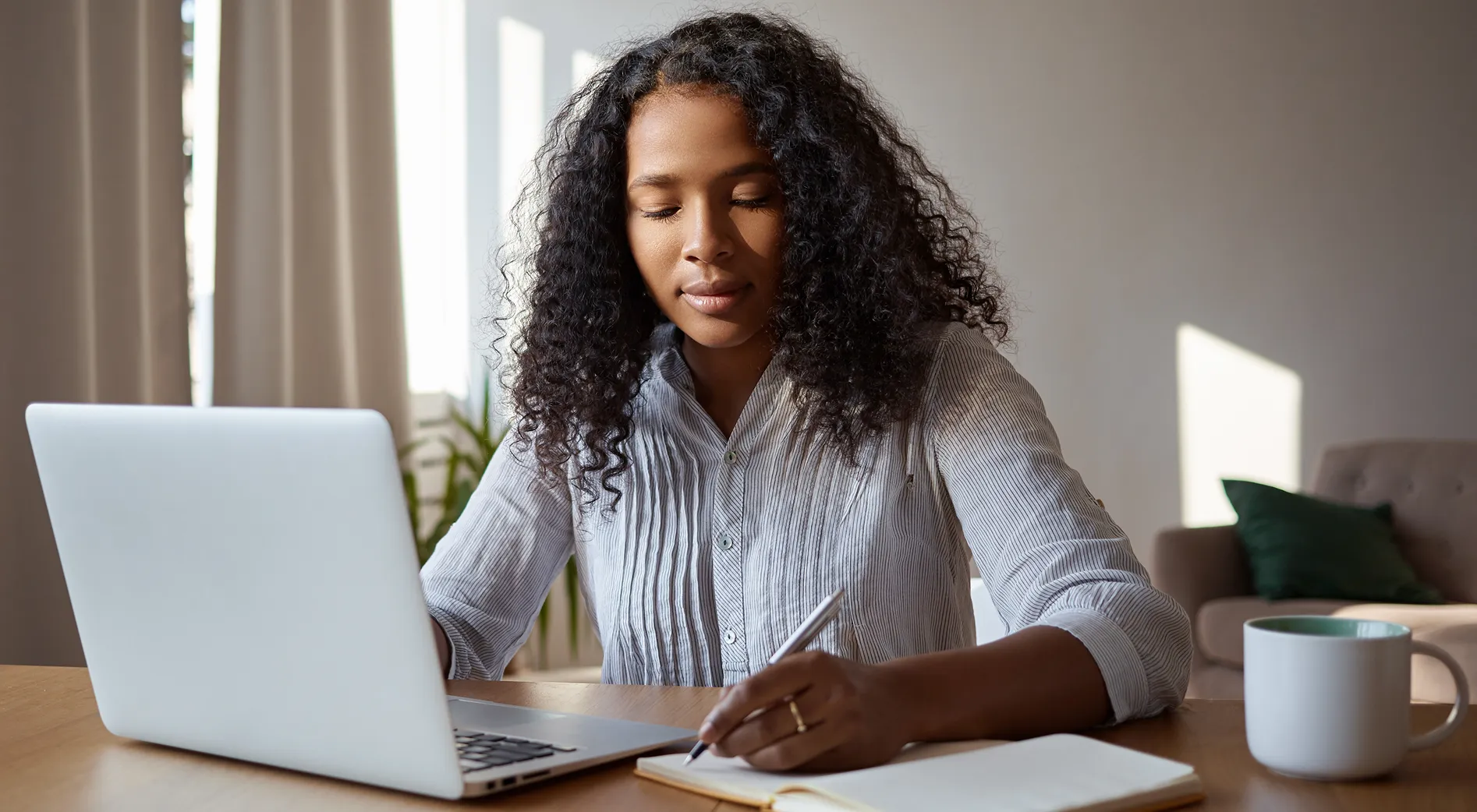 Woman with curly hair works at a laptop, writes in a notebook at a wooden desk with a mug nearby in a bright home office.