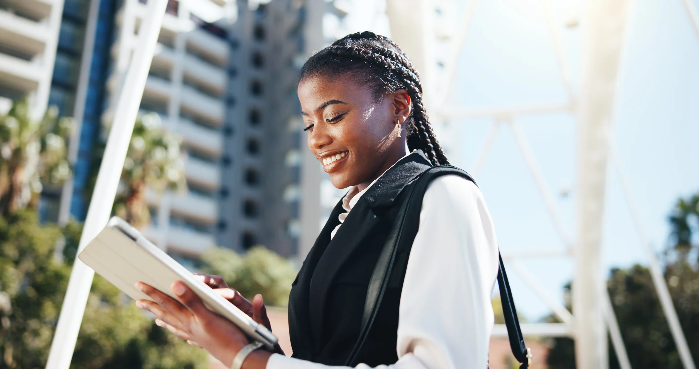 Young Black woman with braided hair, wearing a white blouse and black vest, smiling while reading a tablet outdoors with modern buildings behind her