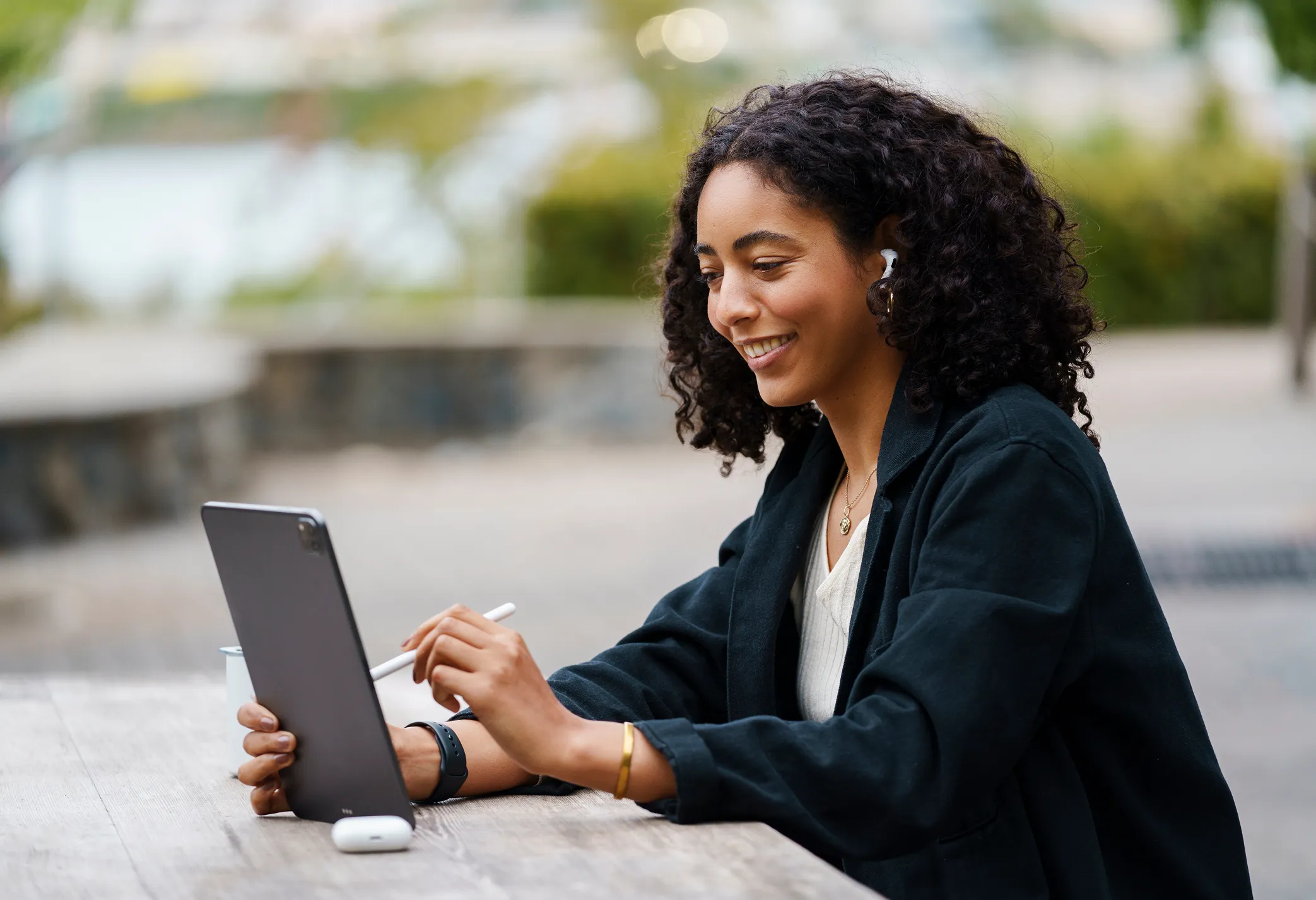 Woman with curly hair wearing earbuds, looking at a tablet while seated at an outdoor table.