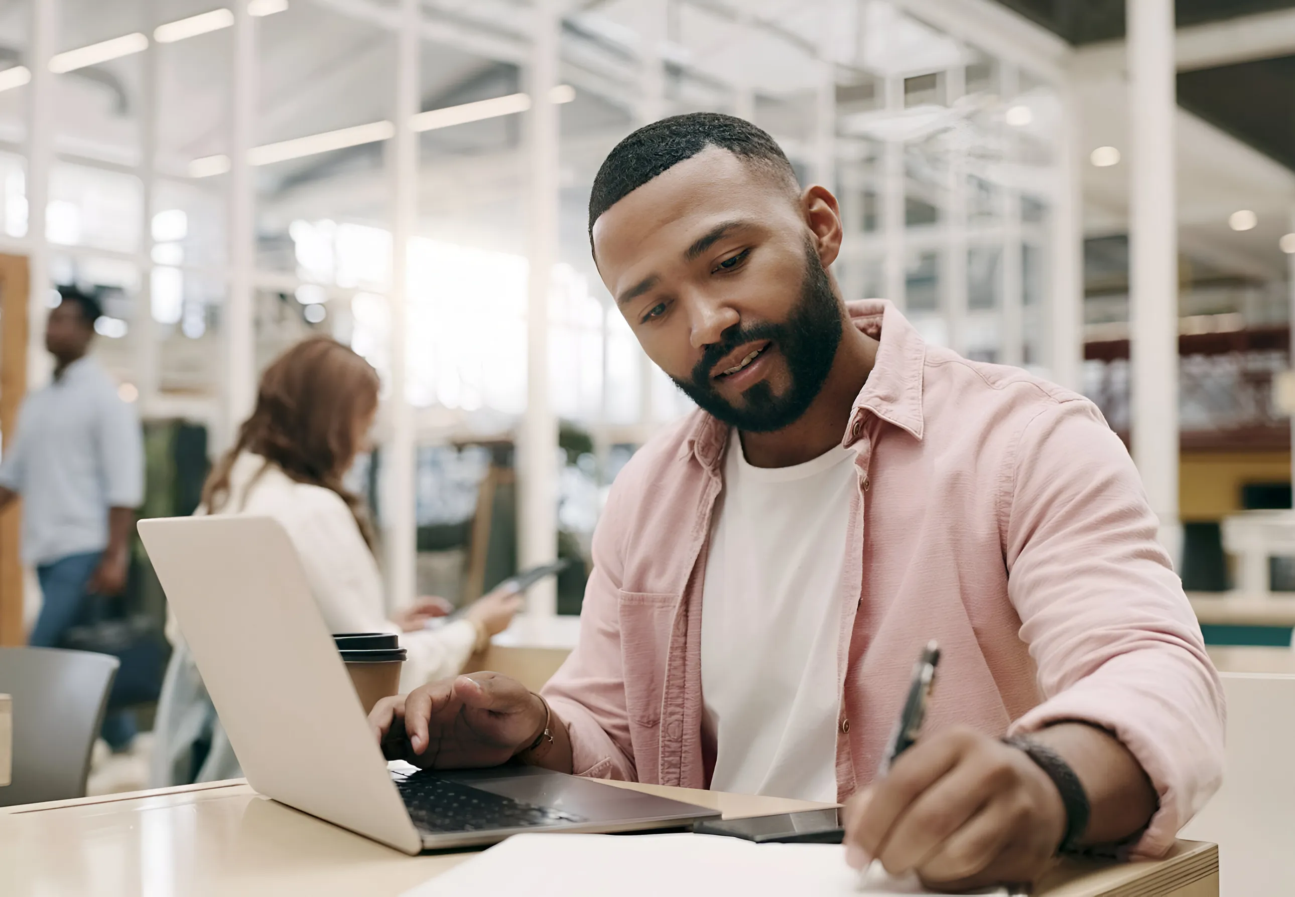 Man in a pink shirt typing on a laptop at a bright cafe or coworking space, coffee cup nearby.