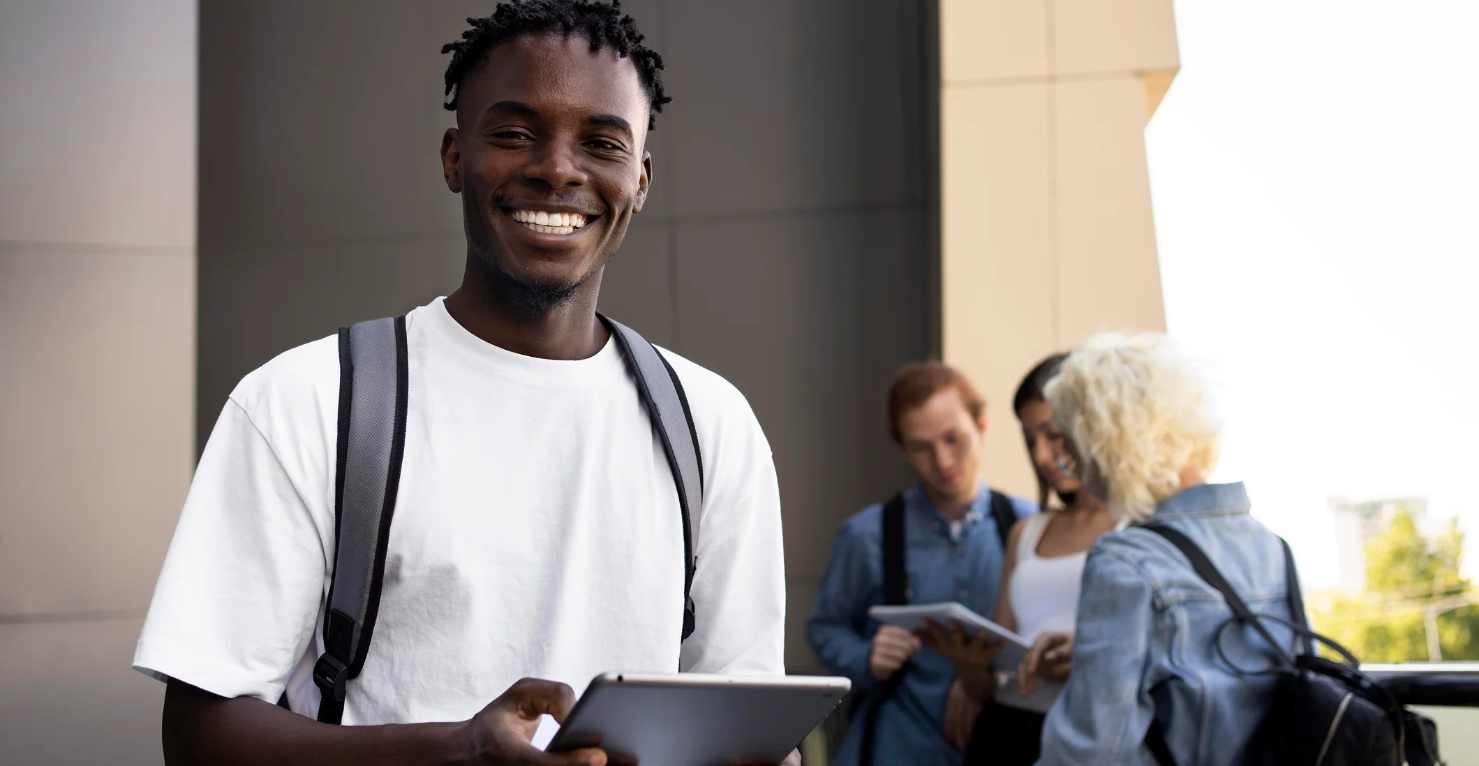 Smiling young man in a white shirt with a backpack holding a tablet; outdoor building in background.