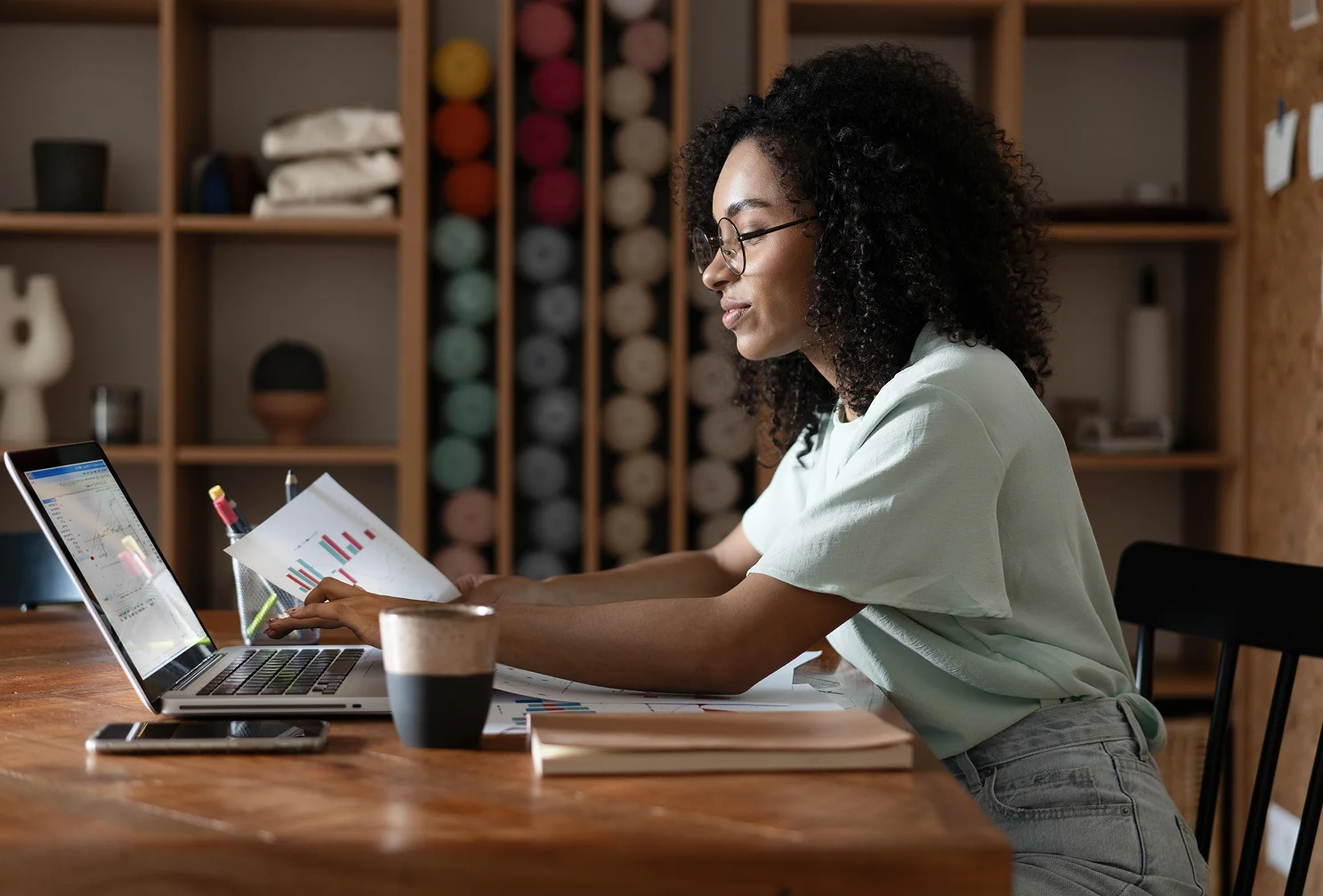 Woman with glasses sits at a wooden table, analyzing charts on paper and data on a laptop, with a coffee cup nearby.