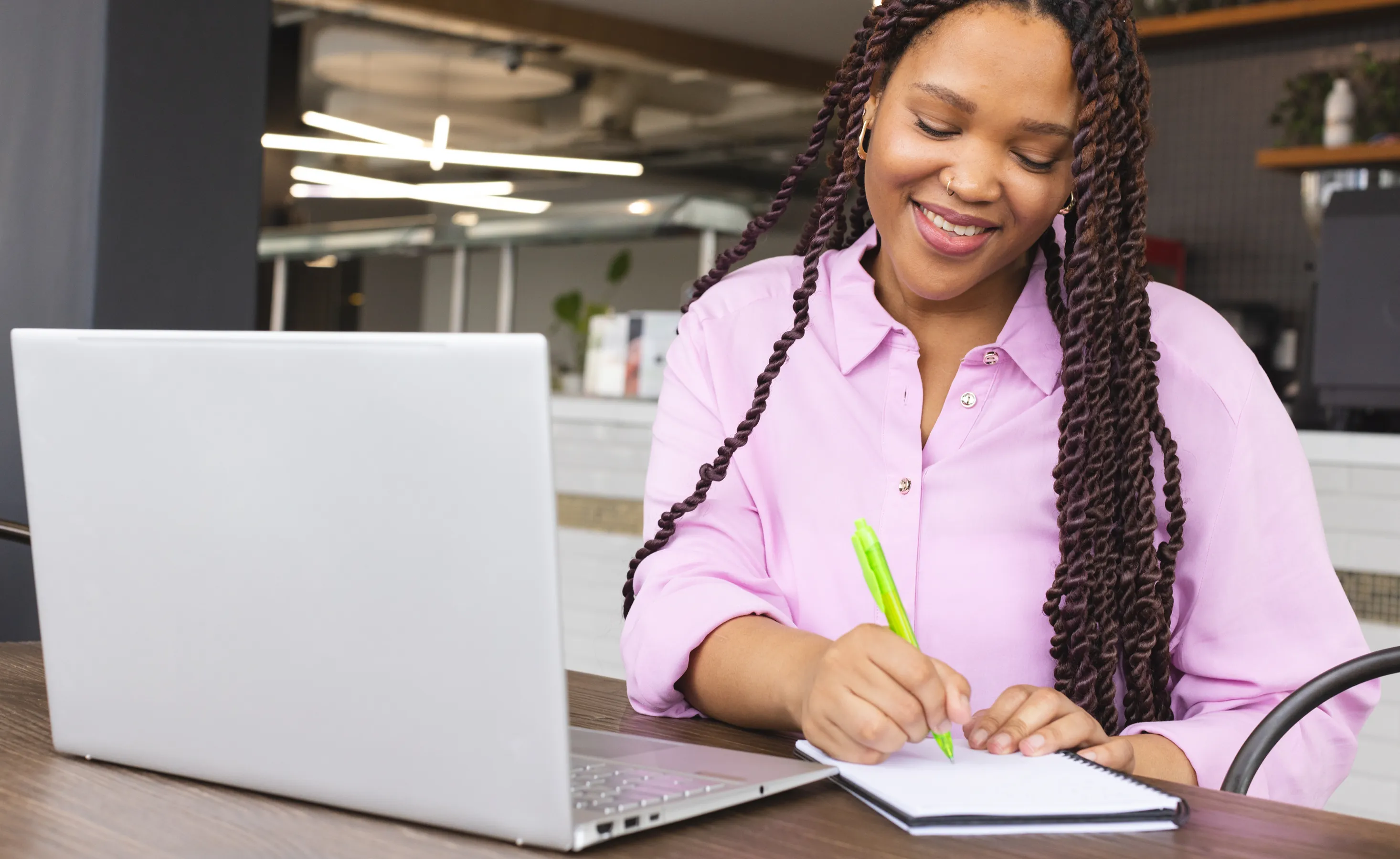 Woman smiling while writing in a notebook beside a silver laptop in a bright office setting, wearing a pink shirt and braids with a green highlighter nearby