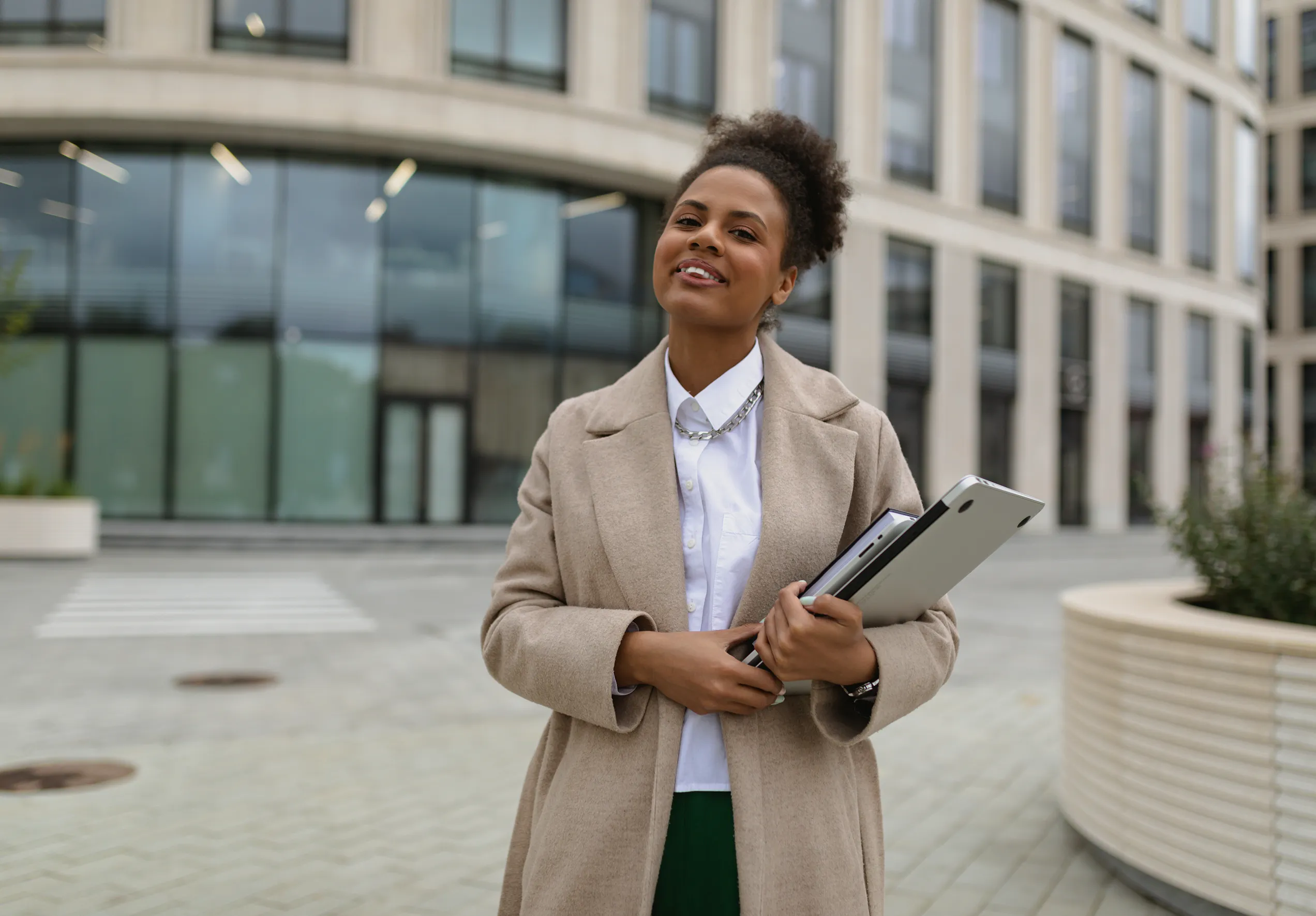 Professional woman in a beige coat holding folders, standing outdoors in front of a modern glass office building.