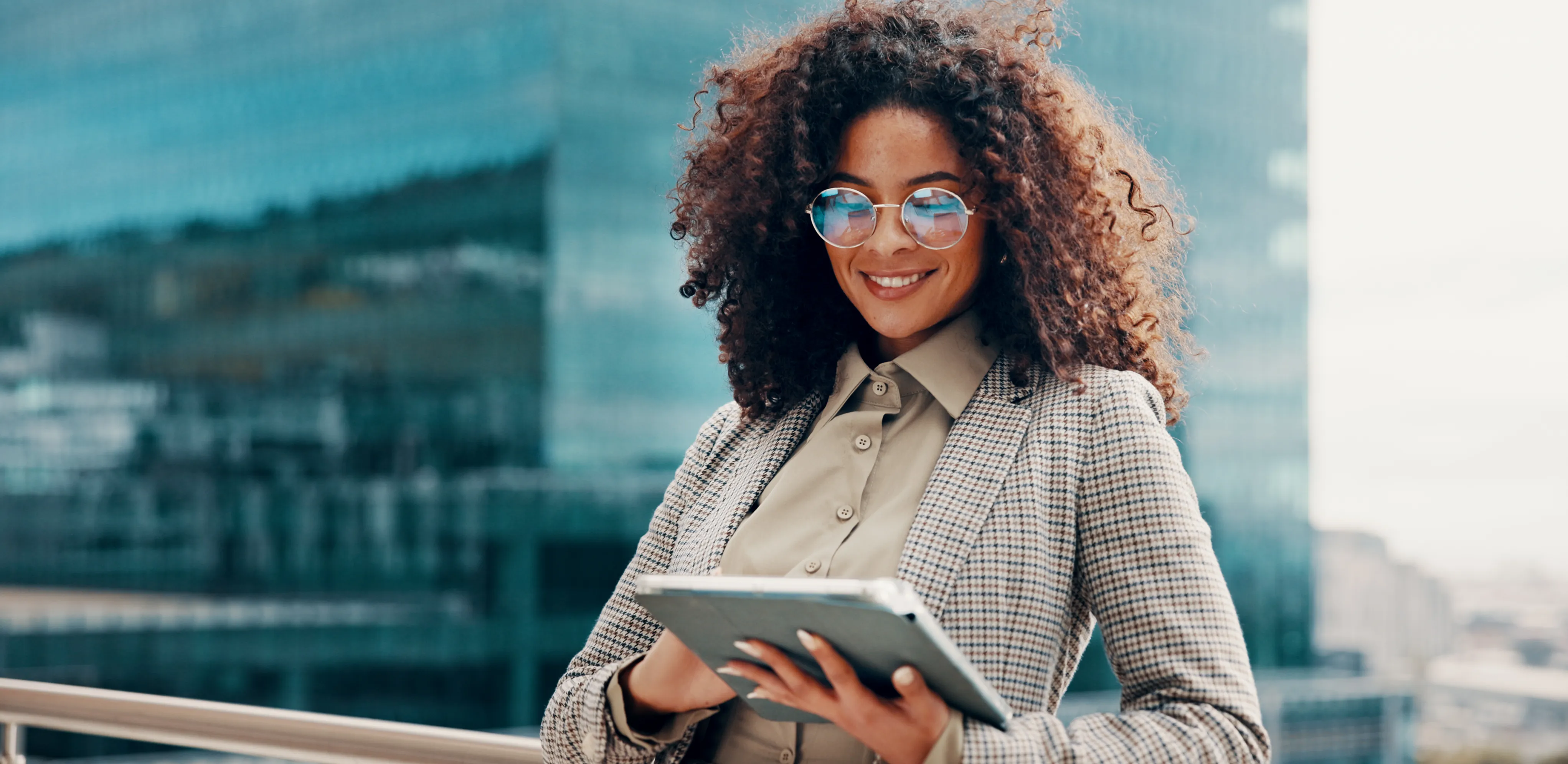Professional woman in a beige blouse and checkered blazer holds a tablet outdoors with city buildings in the background, smiling at the camera.