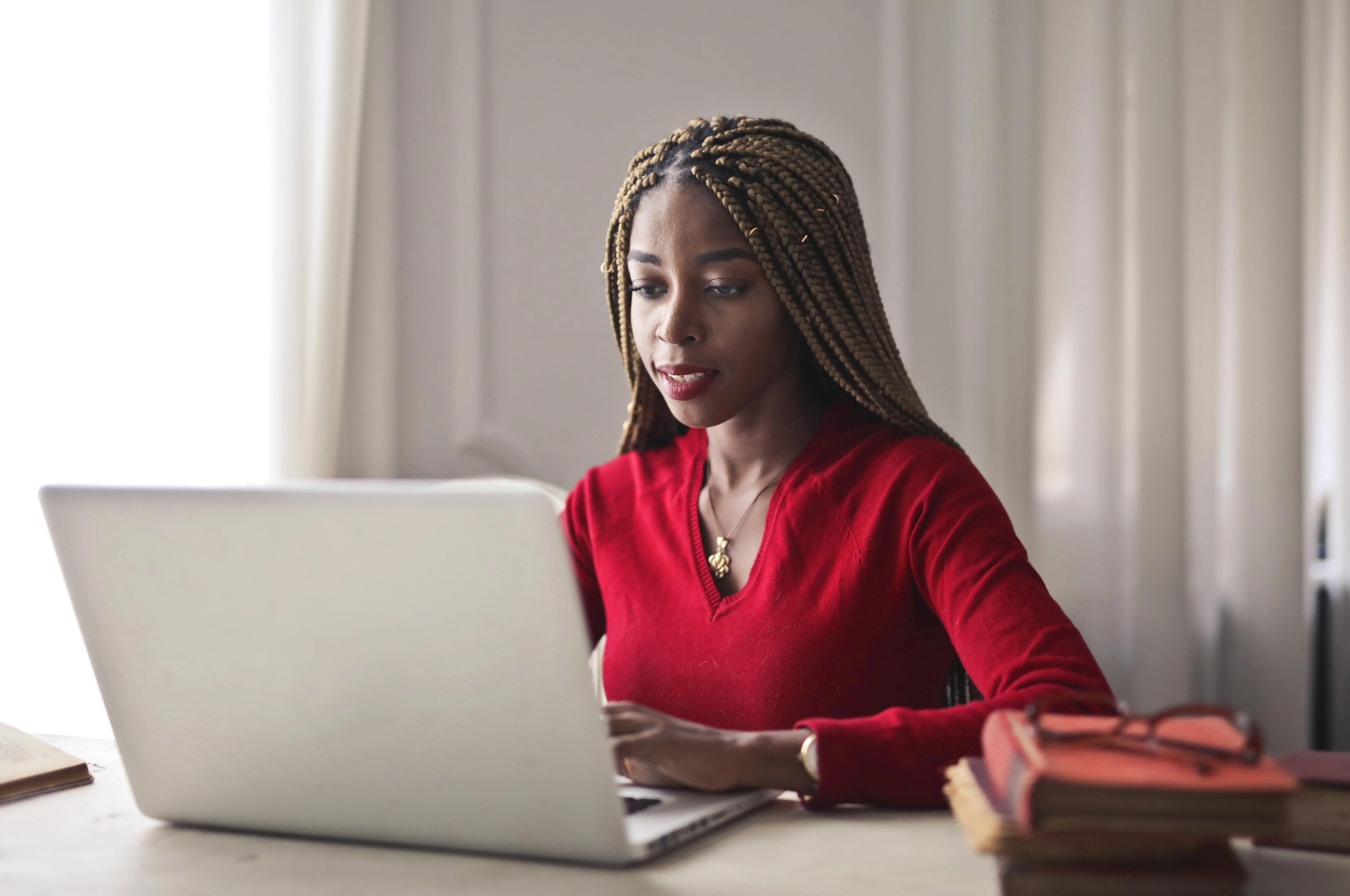 Woman with box braids in a red sweater working on a laptop at a desk.