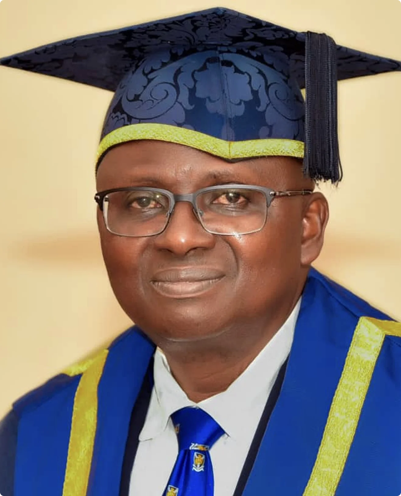 Portrait of a man in a blue graduation cap with yellow trim and gown, wearing glasses and a matching blue tie.
